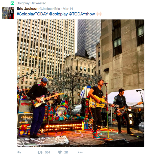 A Twitter post of Coldplay performing on a colorful stage outdoors with tall buildings in the background.