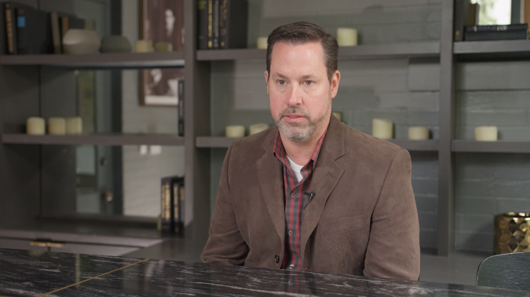 A man seated in an office with books and candles on shelves behind him.