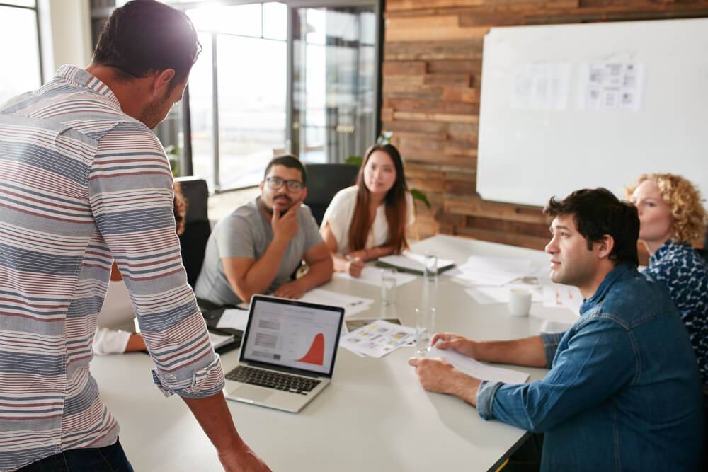 A standing person presenting to a group of colleagues sitting at a conference table with laptops and papers.